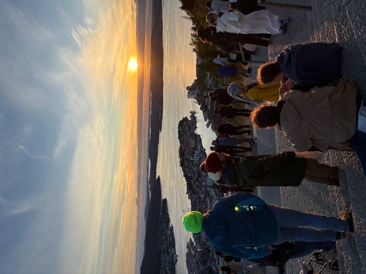 Hikers and sightseers watch a vibrant sunset over Oslofjord from a hilltop viewpoint.