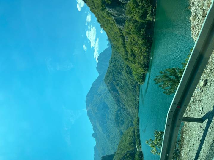 Mountain lake bordered by forested hills under a bright blue sky, viewed from roadside