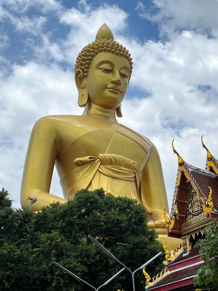 Close-up of a massive golden standing Buddha with ornate temple roof against a partly cloudy sky