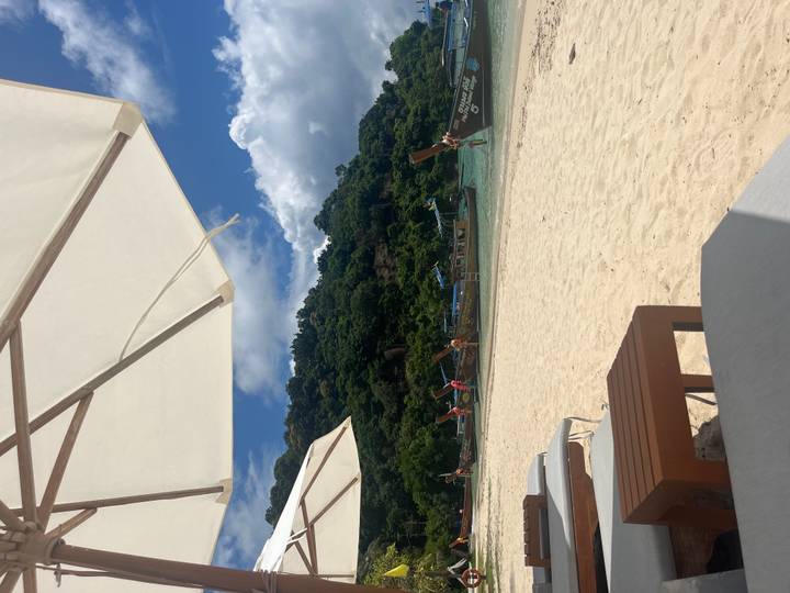 Sandy beach scene with long-tail boats anchored near a forested hillside under sun umbrellas