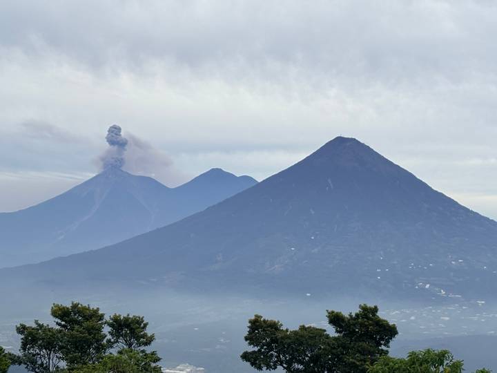 Smoking Fuego volcano erupts beside the conical Acatenango peak against a misty morning sky.