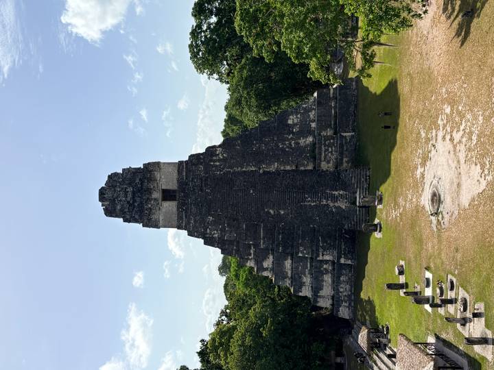 Temple I of Tikal soars above surrounding jungle clearing under bright tropical sky, ancient stones stark against green forest.