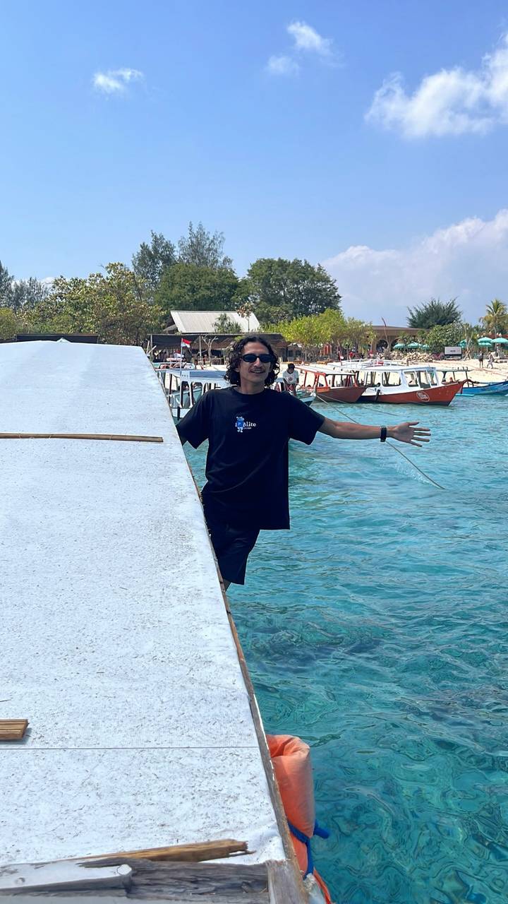 Young man spreads his arms while standing on a boat docked in bright turquoise water with island backdrop.