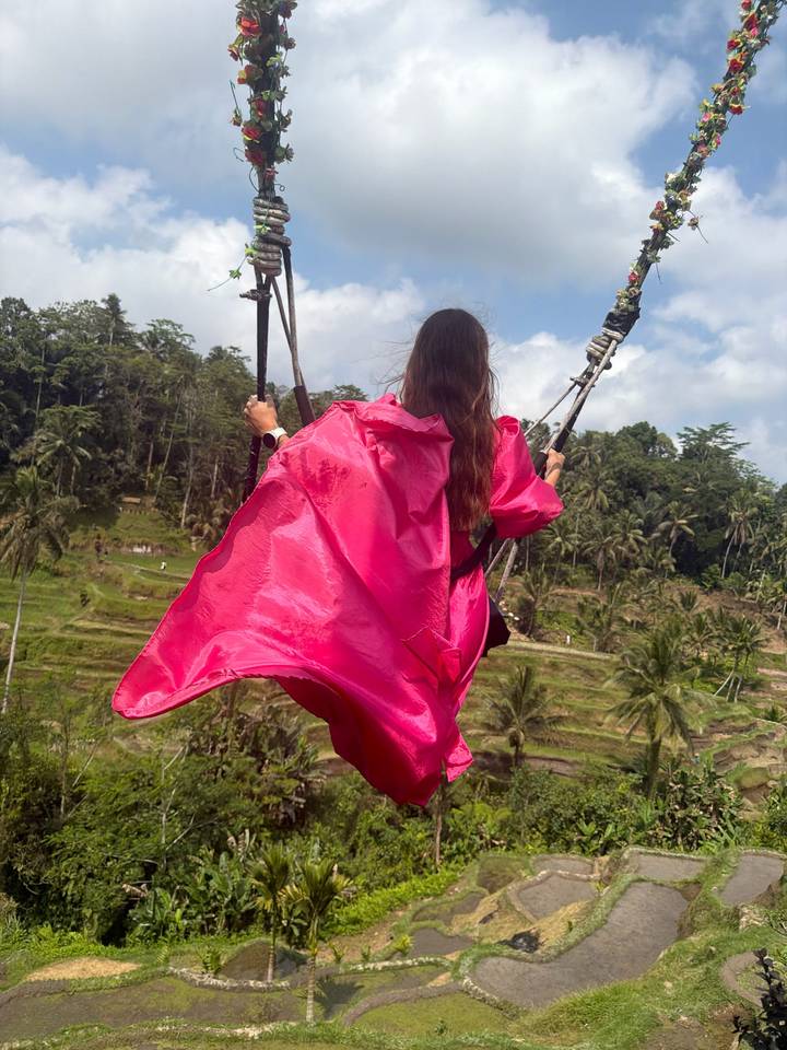 Woman in a flowing pink gown soars on a jungle swing above tiered rice terraces and palm trees.