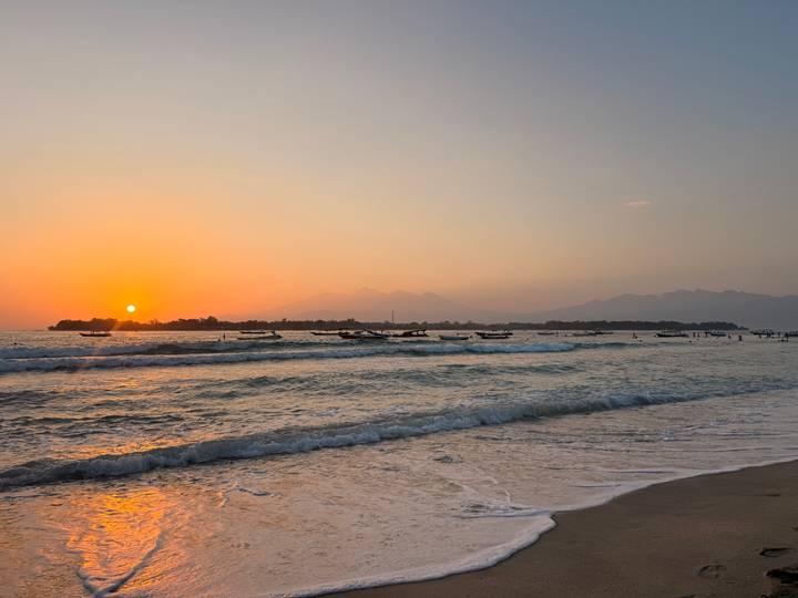 Golden sunset over gentle surf with fishing boats and distant mountains along a tranquil beach.