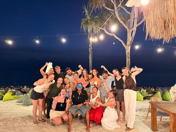 Cheerful group raises fists for a night photo at a beach bar lit by string lights.