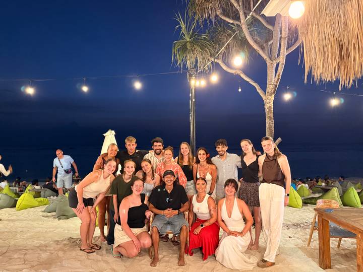Group of travelers pose for an evening portrait on a beach lounge area under hanging bulbs.