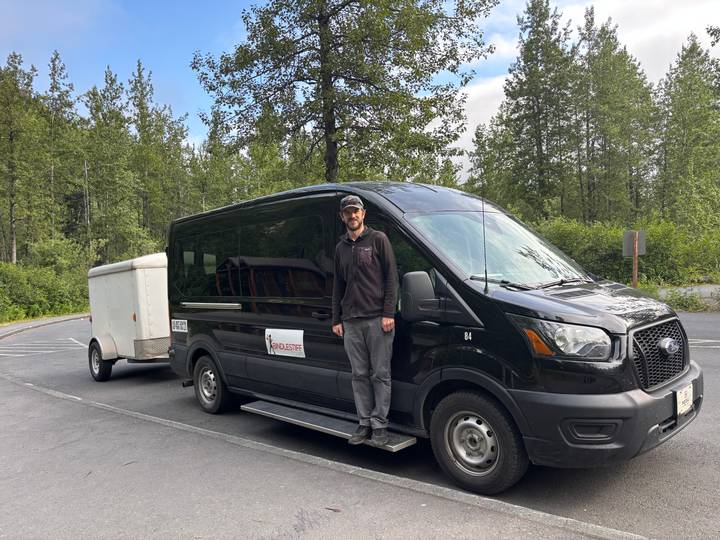 Driver standing beside a black passenger van with a small trailer in a forested parking area
