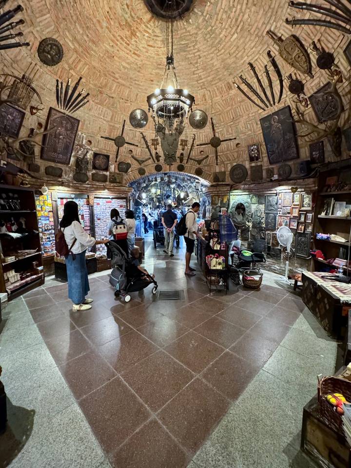 Cave-like souvenir and handicraft shop with travelers browsing shelves and carts under a decorated stone ceiling.