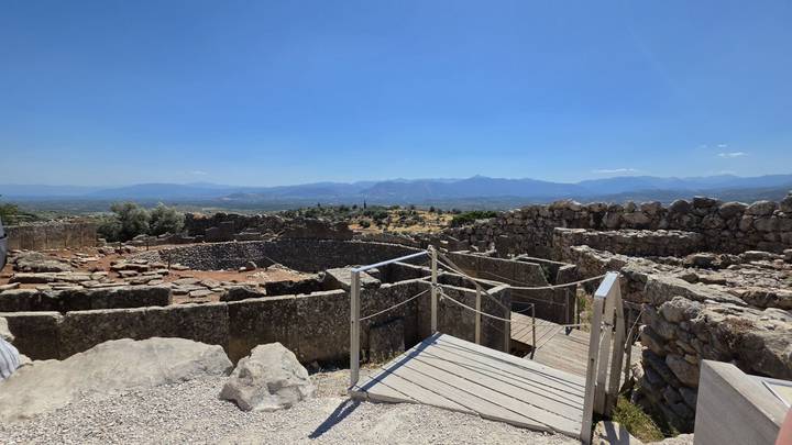 Stone walls and a circular grave complex overlook a valley and distant mountains at an archaeological site.