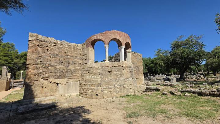 Ruined stone rotunda with brick arches stands among scattered ancient stones and trees.