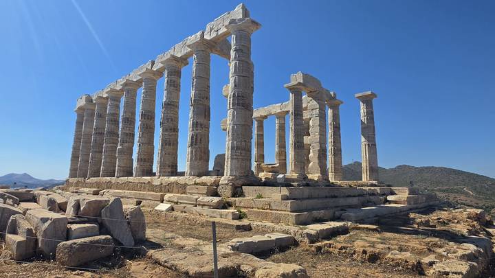 The marble columns of the Temple of Poseidon sit atop a cliff overlooking the sea under a bright blue sky.