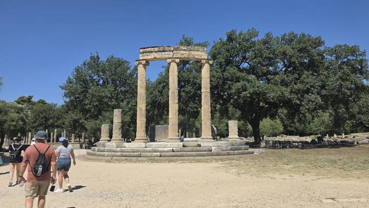 Circular arrangement of ancient columns known as the Philippeion stands in a sandy clearing with tourists nearby.