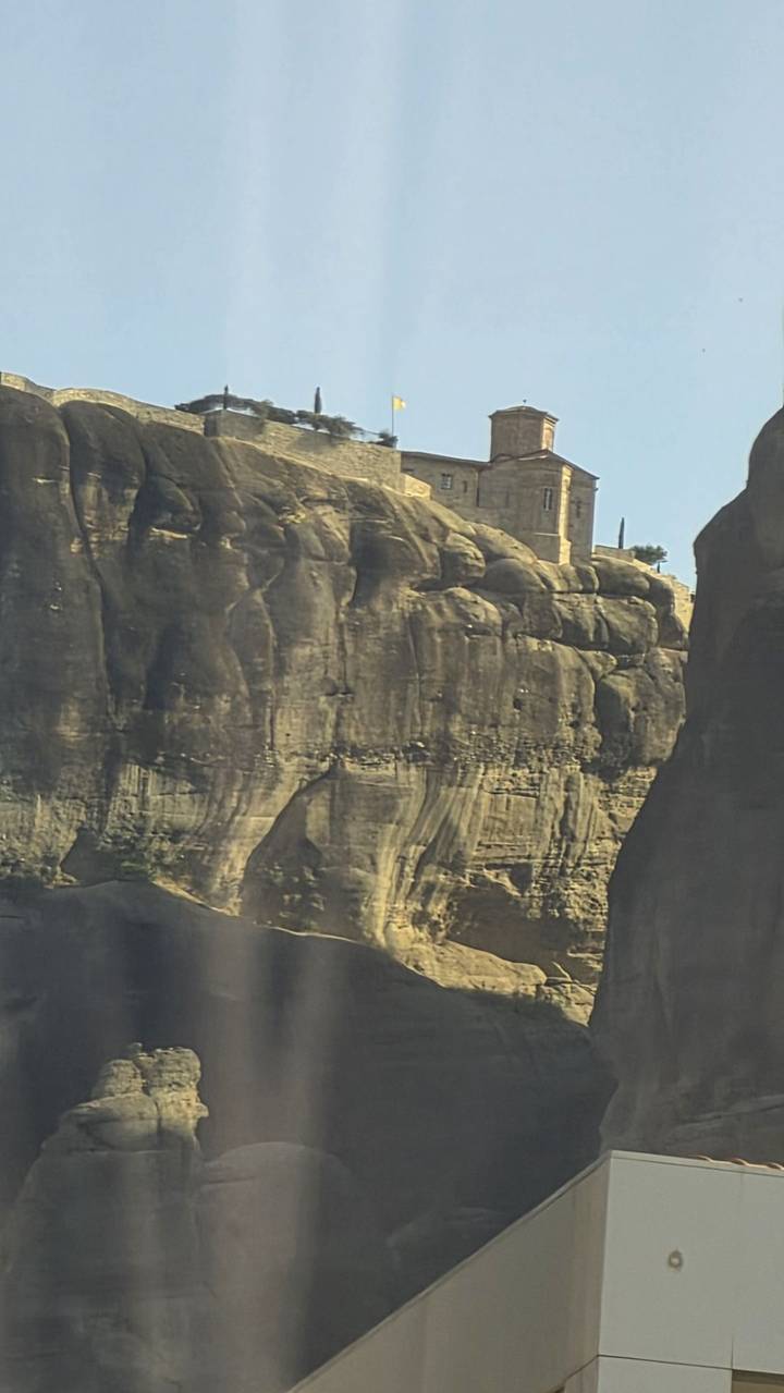Monastery perched atop a towering rock cliff of Meteora seen through a window, slightly out of focus.