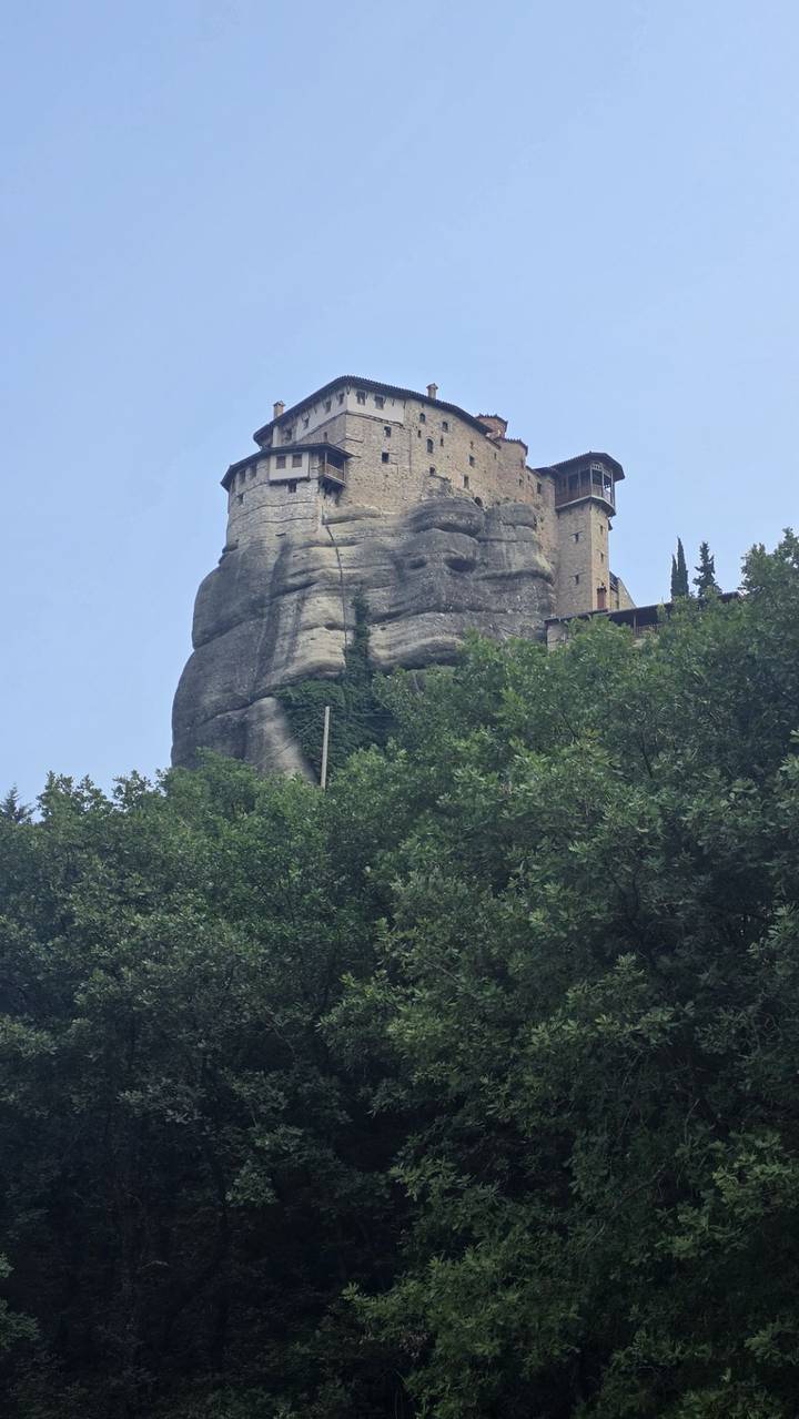 A monastery clings to the side of a tall Meteora rock pillar framed by leafy green trees.
