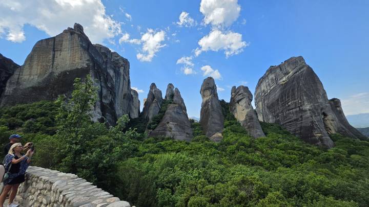 Towering gray sandstone pillars of Meteora rise above lush forest while visitors take photos from a stone wall.