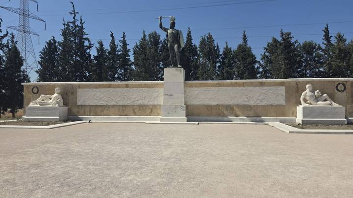 Statue of King Leonidas stands at the Thermopylae monument flanked by marble relief walls and cypress trees.