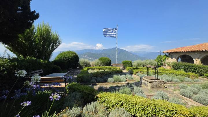 Well-kept monastery garden with Greek flag, trimmed hedges and distant mountain views under blue sky.