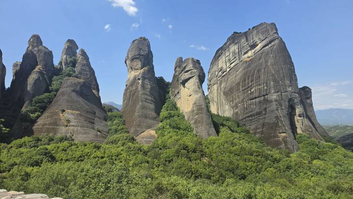 Series of tall, narrow Meteora rock pinnacles rise against a clear blue sky above green forest.