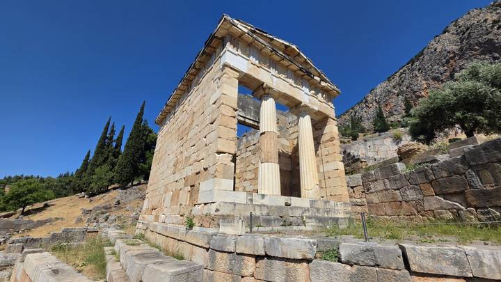 Small marble Treasury of the Athenians stands against a rocky hillside and cypress trees at Delphi.