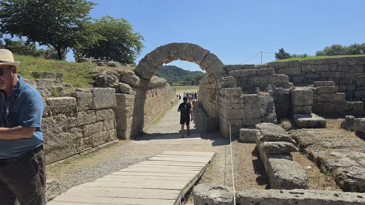 Stone archway leads visitors into the ancient Olympic stadium with grassy field beyond.