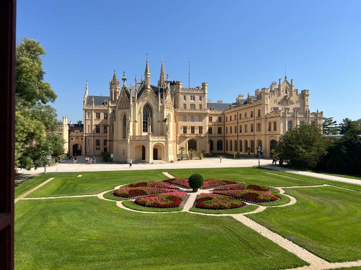 Elegant neo-gothic chateau with ornate spires overlooking a manicured lawn and circular flower bed under a clear blue sky
