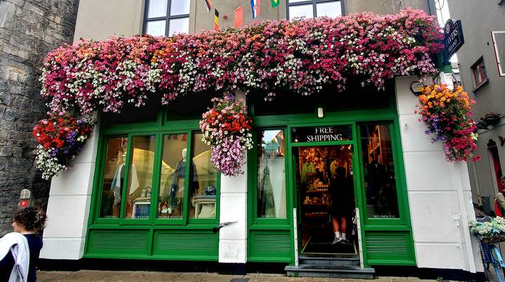Charming storefront with bright green woodwork and cascades of colorful hanging flowers, sign reads 'Free Shipping'