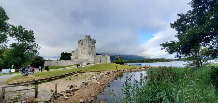 Stone lakeside castle ruin with visitors walking nearby, backed by misty hills and a partly cloudy sky