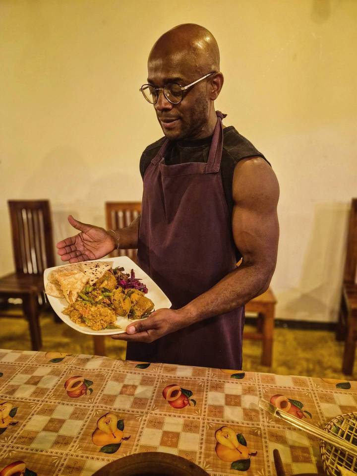 Server presenting a plate of colorful Sri Lankan dishes while wearing an apron inside a rustic eatery.