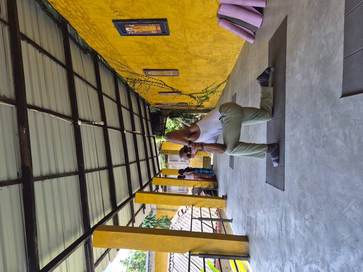 Practitioner holding a yoga pose on a mat in a shaded open corridor with yellow pillars.