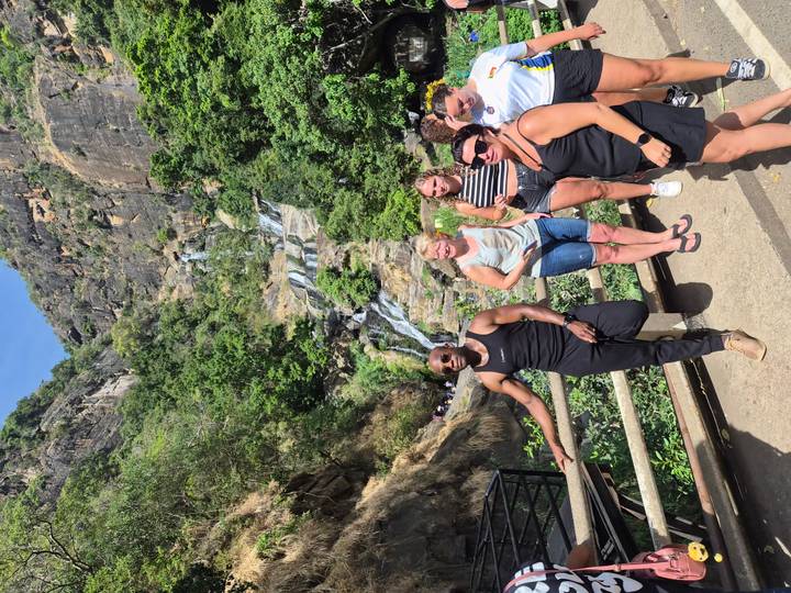 Small tour group posing on a bridge with a multi-tiered jungle waterfall cascading down the cliff behind.