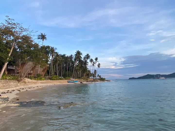 Tropical beach with white sand, swaying palm trees, rustic cottages and a small blue boat on tranquil turquoise water at dusk.