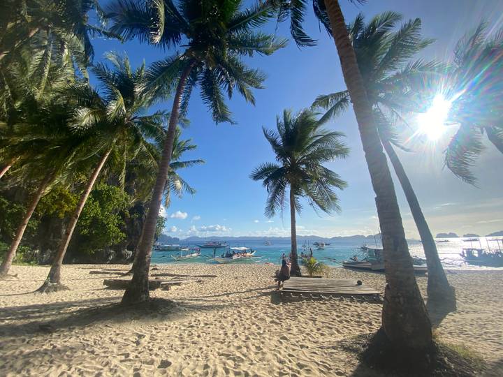 Sunny palm-fringed beach with visitors relaxing on benches while bangka boats bob on the clear blue sea.