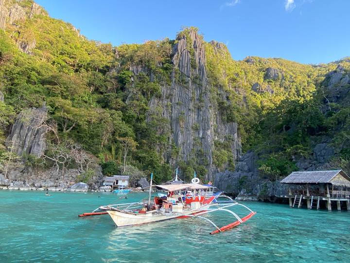 Traditional outrigger boat anchored in crystal-clear lagoon beside towering limestone cliffs covered in green foliage.