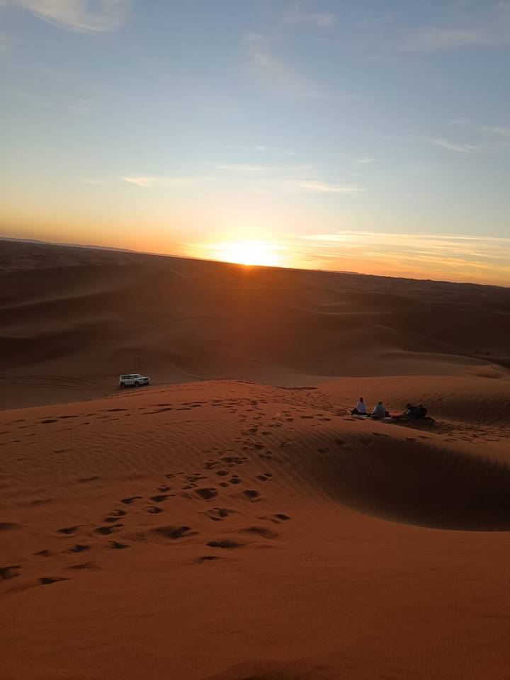 Small group watching a fiery desert sunset from high sand dunes with a lone 4x4 parked below.