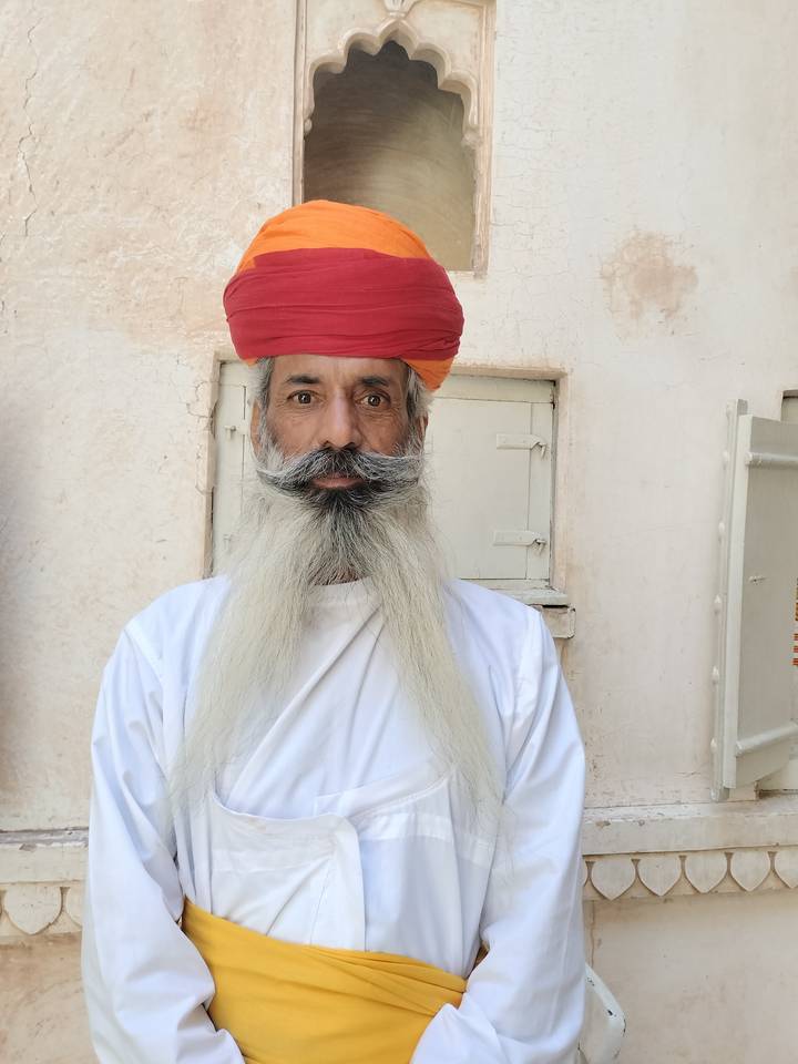 Portrait of an elderly Rajasthani man with long white beard and colorful turban standing against a pale wall.