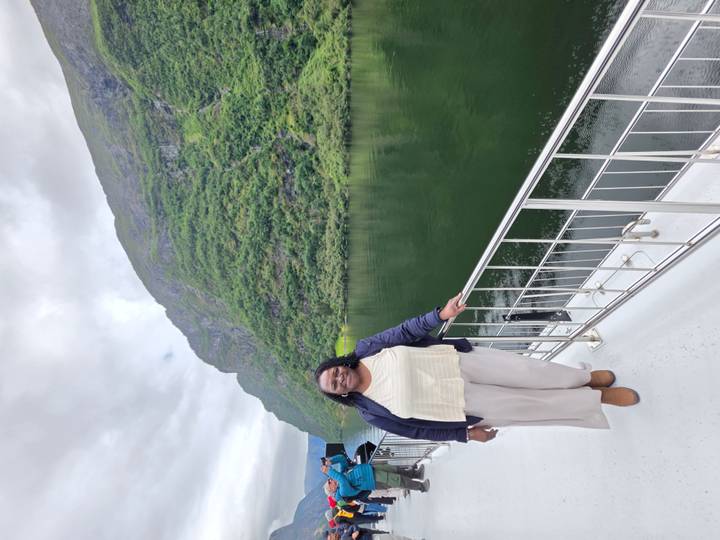 Woman standing on a boat deck with lush green fjord cliffs rising behind her under cloudy skies.