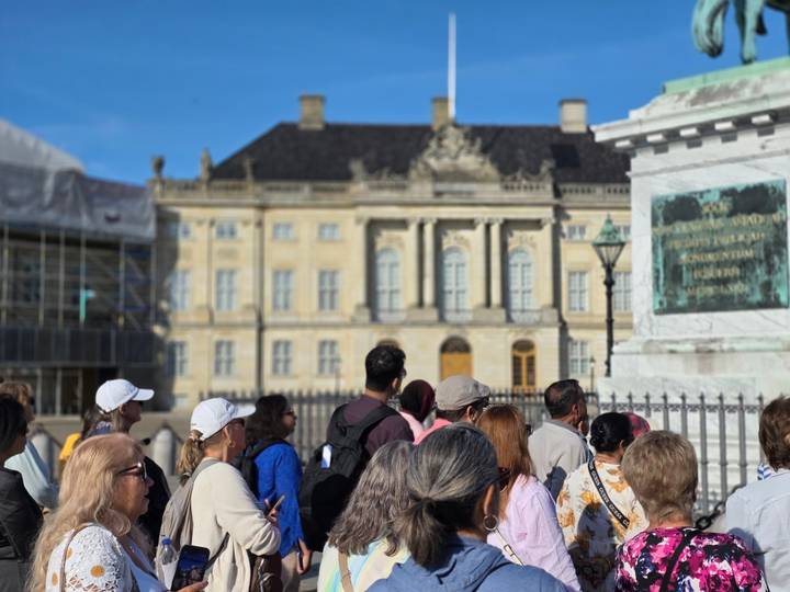 Tour group listens to a guide in front of an elegant palace courtyard under a bright blue sky.