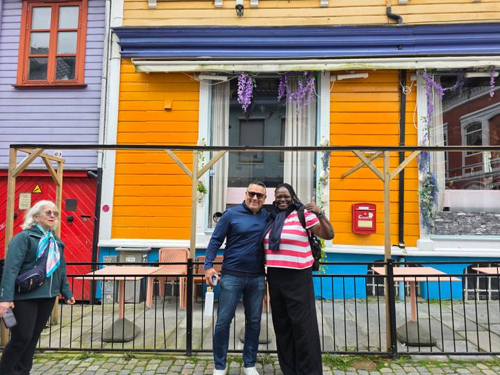 Travelers smile and pose outside a brightly painted wooden façade decorated with flowers.