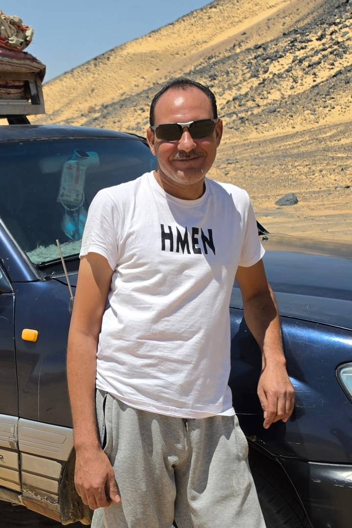 Man in white T-shirt stands by a 4×4 vehicle on a sandy desert track.