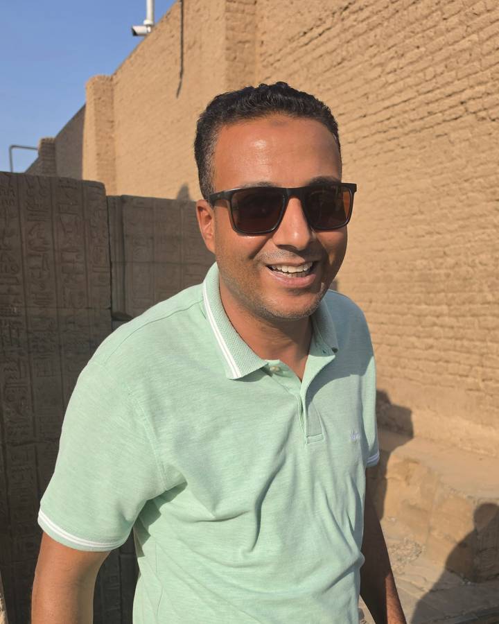 Tour guide wearing sunglasses smiles beside carved hieroglyphic blocks at an ancient temple.