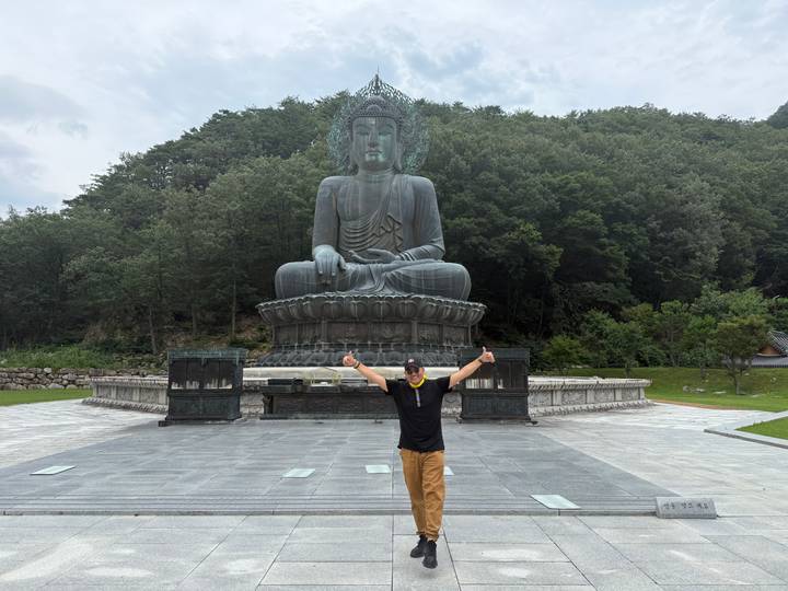 Man posing thumbs up before large seated Buddha statue in forested temple grounds