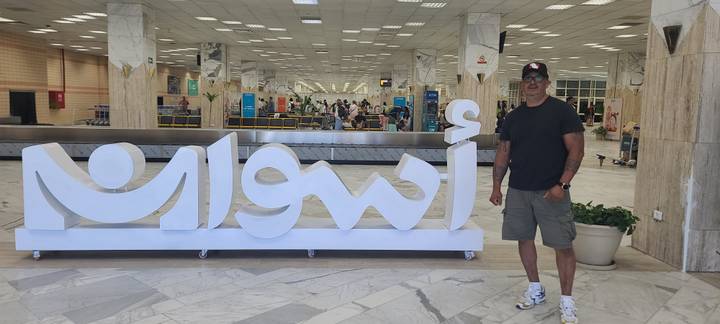 Traveler standing beside large illuminated Aswan sign inside the airport terminal
