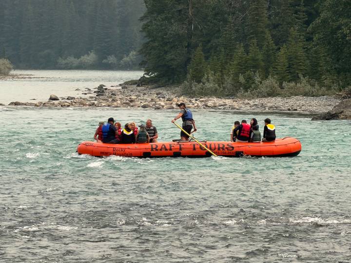 Tourists paddle an orange raft on a glacial river surrounded by evergreen forest