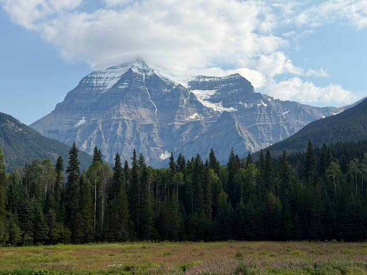 Snow-capped Mount Robson rises dramatically above dense pine forests under a partly cloudy sky
