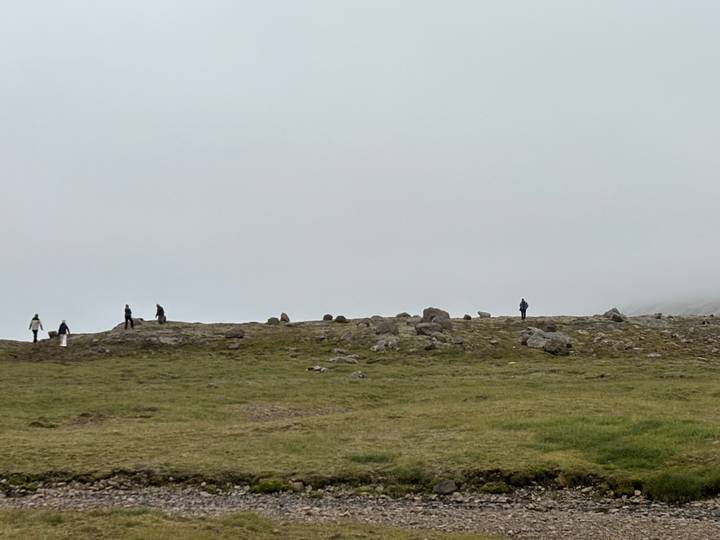 Tiny silhouettes of hikers spread across a foggy, grassy ridge under low clouds.
