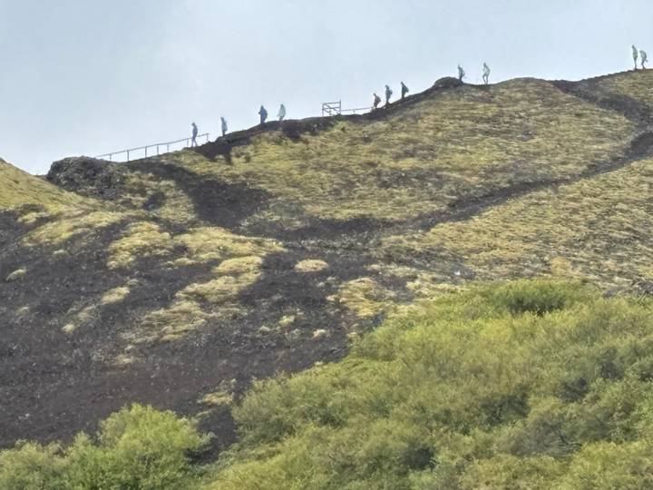 Line of hikers ascending the ridge of a mossy volcanic slope with switchback paths.