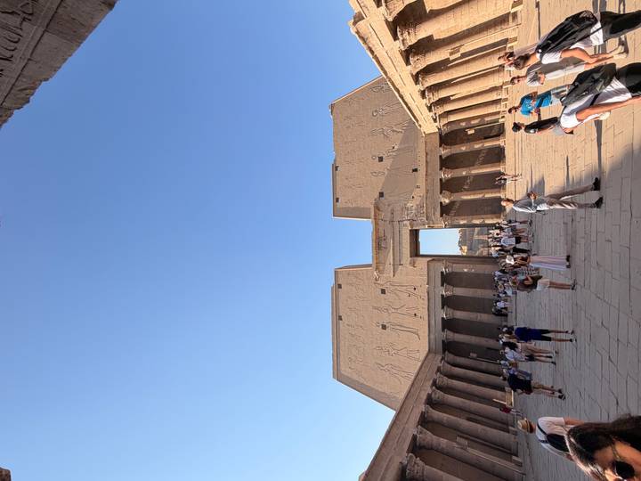 Spacious courtyard and pylon entrance of Edfu Temple with tourists exploring under blue sky.