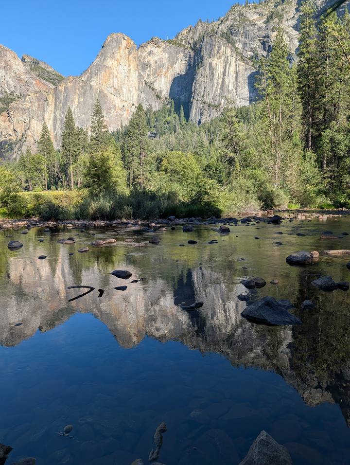 Clear shallow river with scattered rocks reflecting tall pines and granite cliffs.