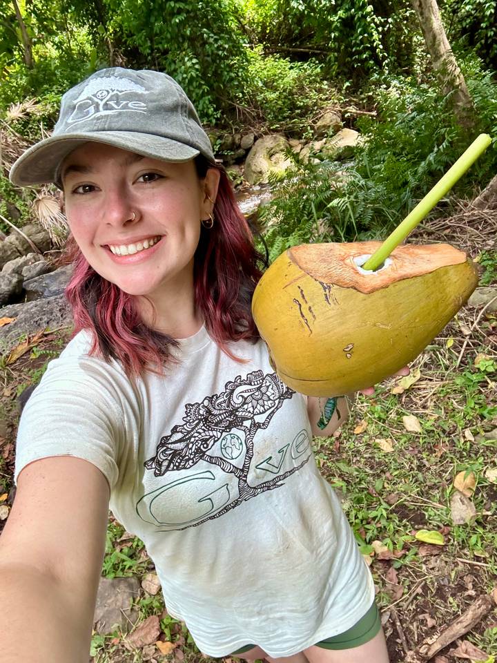 Smiling traveler taking a selfie while holding a freshly opened coconut with a straw in lush surroundings.